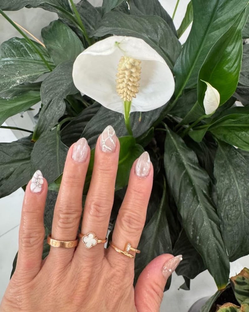 Sheer iridescent nails with white 3D floral and pearl accents, held against a peace lily